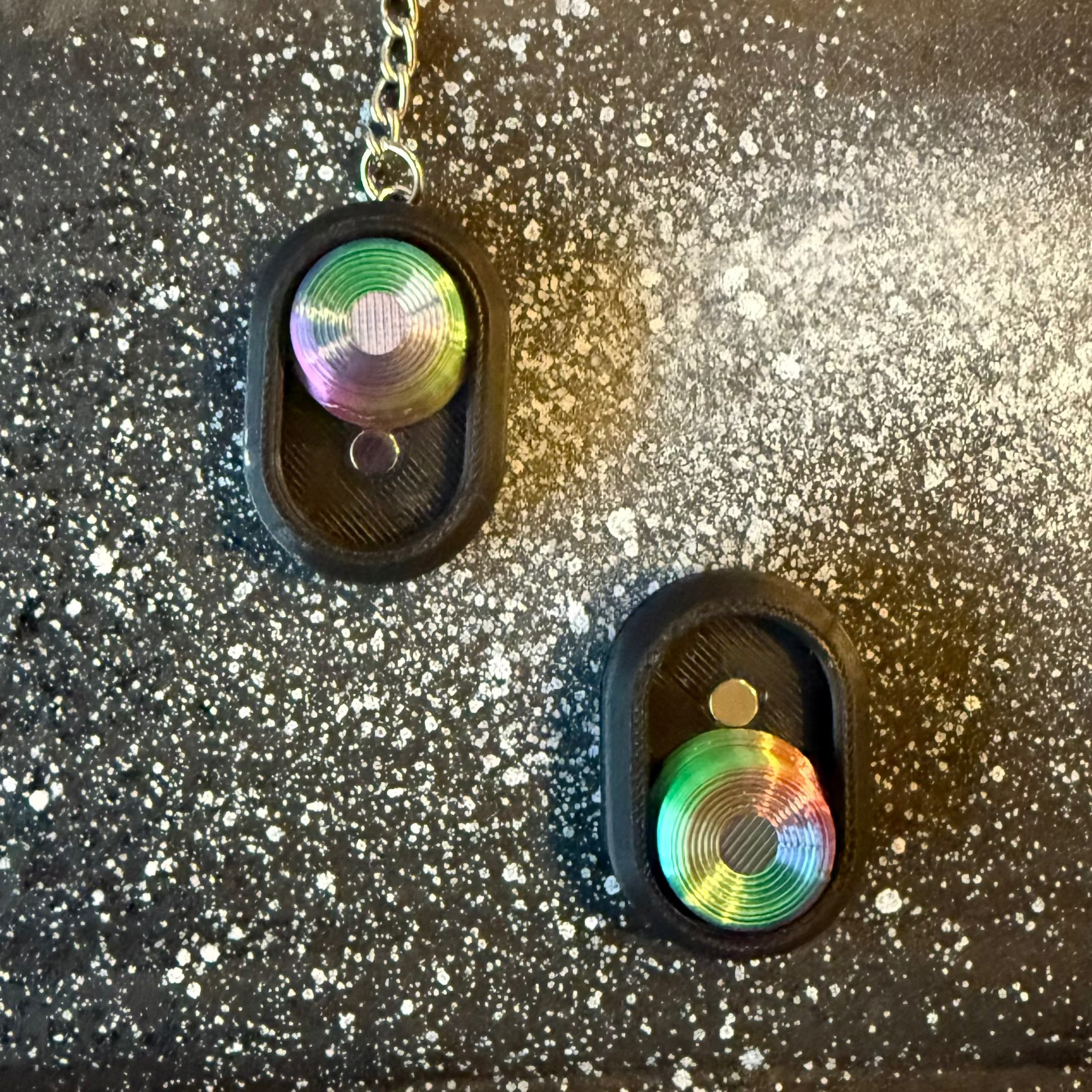 Two Snappy™ Northern Lights Keychains with aurora-colored tops and black oval bases on a starry black background. Executive toy for sensory play.